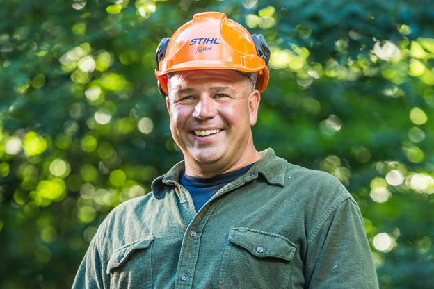 Portrait of a smiling male employee wearing an orange STIHL hard hat and hearing protection in a forest setting.